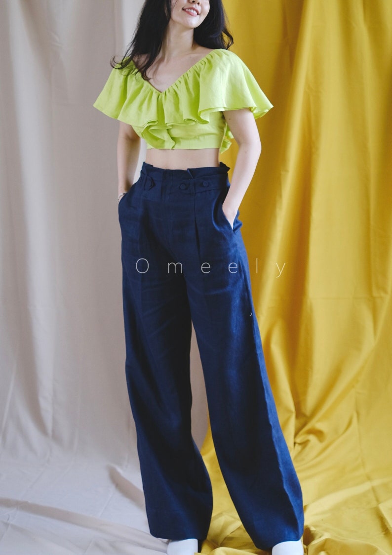 May include: A woman is wearing a lime green ruffled crop top and dark blue wide-leg pants. The pants have a high waist and a belt. The woman is standing in front of a yellow and beige background.
