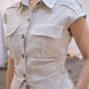 May include: A beige short-sleeved button-down shirt featuring a classic collar, two chest pockets with flaps, and dark brown buttons. The shirt has a tailored fit with a slightly curved hemline and includes the text Omeely printed on the front.