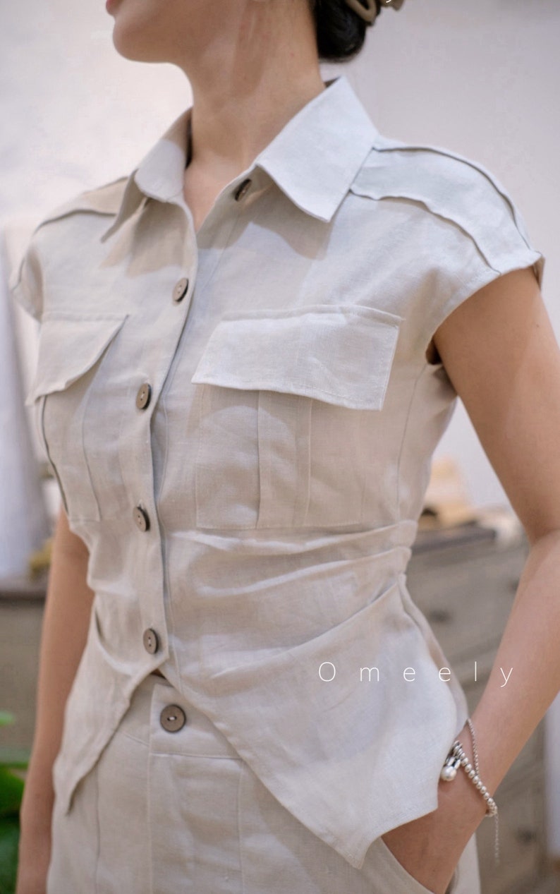 May include: A beige short-sleeved button-down shirt featuring a classic collar, two chest pockets with flaps, and dark brown buttons. The shirt has a tailored fit with a slightly curved hemline and includes the text Omeely printed on the front.
