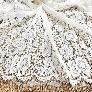 May include: Close-up of white lace fabric with a floral pattern. The delicate lace is draped, showcasing its intricate design and texture. The fabric's edges have a scalloped trim, adding to its elegance. Ideal for apparel and craft projects.