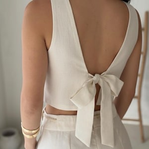 May include: Cream-colored sleeveless top with a deep V-neck and a bow detail at the back. The top is paired with matching high-waisted pants. Gold bracelets adorn the arm. The outfit is made of a textured fabric, suggesting linen or a similar material.