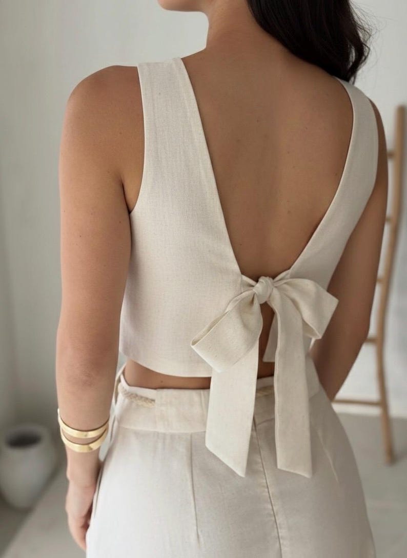 May include: Cream-colored sleeveless top with a deep V-neck and a bow detail at the back. The top is paired with matching high-waisted pants. Gold bracelets adorn the arm. The outfit is made of a textured fabric, suggesting linen or a similar material.