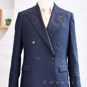 Op de afbeelding: Een marineblauwe double-breasted blazer met puntrevers, vier donkere knopen en een rood stiksel op de linker revers. De blazer wordt gedragen over een lichtgekleurd overhemd met kraag. De tekst Omeely staat in het wit op de voorzijde.