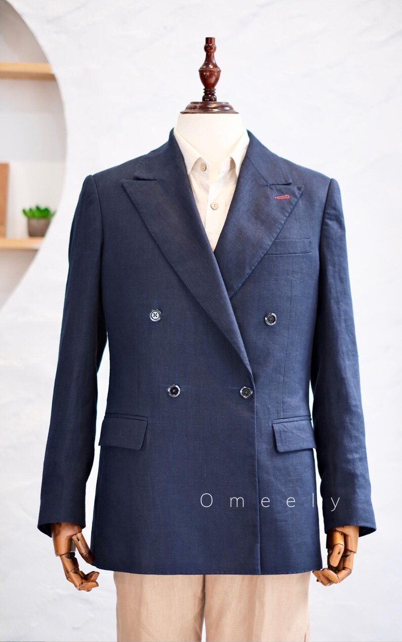 Op de afbeelding: Een marineblauwe double-breasted blazer met puntrevers, vier donkere knopen en een rood stiksel op de linker revers. De blazer wordt gedragen over een lichtgekleurd overhemd met kraag. De tekst Omeely staat in het wit op de voorzijde.
