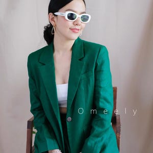 May include: A woman wearing a green blazer and matching trousers. She is wearing white sunglasses and has her hair pulled back. The text "Omely" is visible in the image.