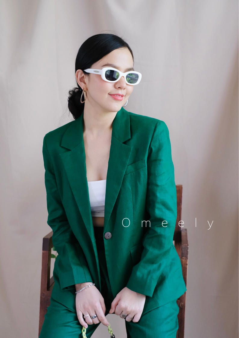 May include: A woman wearing a green blazer and matching trousers. She is wearing white sunglasses and has her hair pulled back. The text "Omely" is visible in the image.