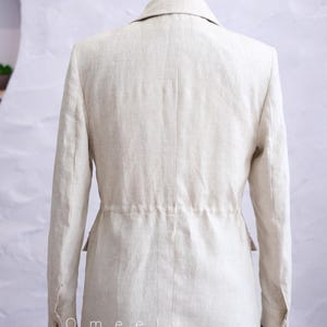May include: A beige blazer with long sleeves, a collared neckline, and a cinched waist featuring a drawstring detail. The garment has buttoned cuffs and two side pockets. The text Omeely is printed in white on the lower left side of the blazer.