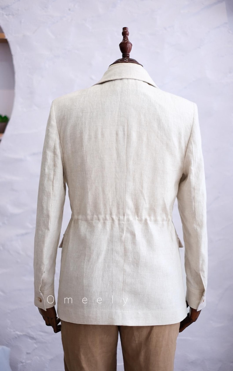 May include: A beige blazer with long sleeves, a collared neckline, and a cinched waist featuring a drawstring detail. The garment has buttoned cuffs and two side pockets. The text Omeely is printed in white on the lower left side of the blazer.