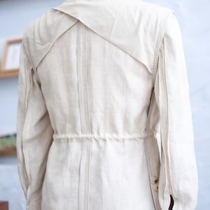 May include: An unlined beige jacket featuring a collared neckline, long sleeves with buttoned cuffs, and a drawstring waist. The back design includes a layered yoke detail with vertical stitching running down the center, creating a structured look.