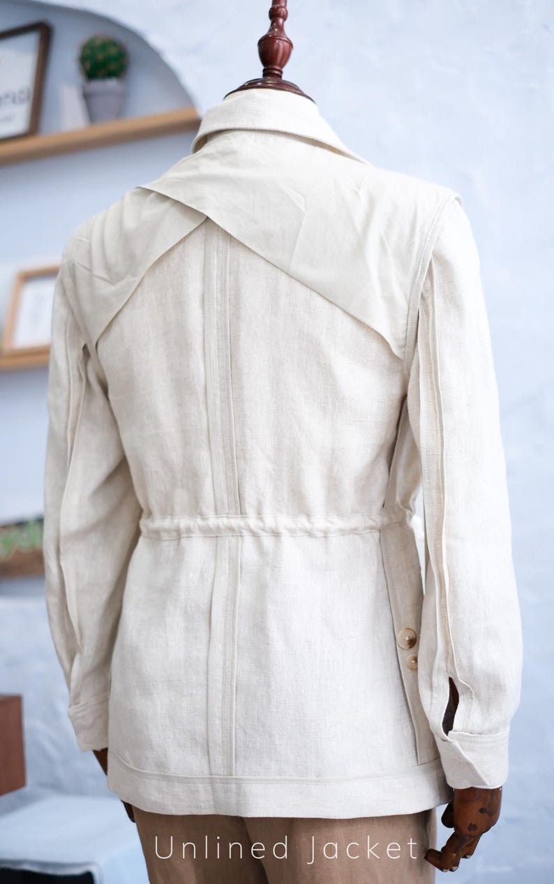 May include: An unlined beige jacket featuring a collared neckline, long sleeves with buttoned cuffs, and a drawstring waist. The back design includes a layered yoke detail with vertical stitching running down the center, creating a structured look.