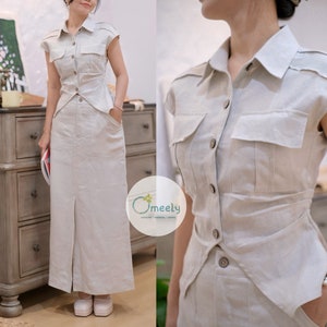 May include: A beige two-piece set consisting of a short-sleeved button-down shirt with two chest pockets and a matching floor-length skirt with a front slit and side pockets. The shirt features a pointed collar and a tapered waistline design.