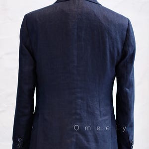 Op de afbeelding: Een donkerblauwe blazer op een paspop, voorzien van een klassieke kraag, lange mouwen met knoopmanchetten en een centrale naad op de rug. Het woord Omeely is in kleine witte letters gedrukt op de rechteronderkant van het achterpaneel.