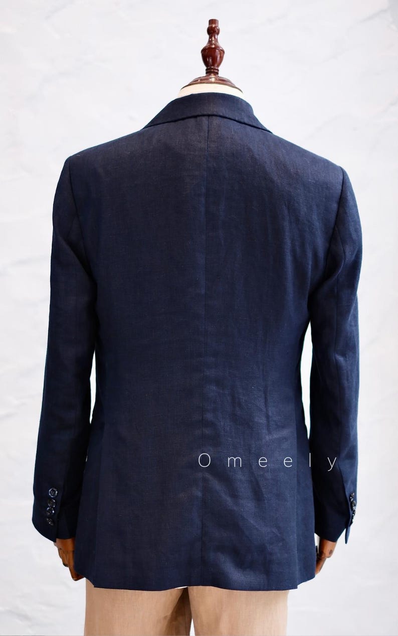 Op de afbeelding: Een donkerblauwe blazer op een paspop, voorzien van een klassieke kraag, lange mouwen met knoopmanchetten en een centrale naad op de rug. Het woord Omeely is in kleine witte letters gedrukt op de rechteronderkant van het achterpaneel.