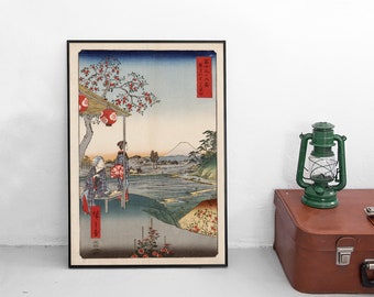 Op de afbeelding: Een ingelijste ukiyo-e houtsnede met een landschap met de berg Fuji in de verte, twee figuren onder een boom met rode lantaarns en rode tekstcartouches. Het kunstwerk is ingelijst in een dunne zwarte lijst met een crèmekleurige rand.