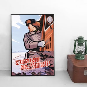 Puede incluir: Un póster enmarcado con una ilustración estilizada de una persona con gorro de piel y abrigo asomándose desde un vehículo. La obra incluye un paisaje nevado y el texto coreano en rojo: "토지정리사업에 계속 큰 힘을 넣자!"