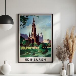May include: A framed travel poster print featuring an illustration of the Scott Monument in Edinburgh, Scotland. The artwork displays the dark gothic spire rising above green parkland and trees under a blue sky, with the text THE SCOTT MONUMENT EDINBURGH.