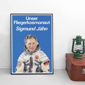 Puede incluir: Un póster enmarcado con fondo azul y un retrato de un hombre con un traje espacial blanco. El póster dice "Unser Fliegerkosmonaut Sigmund Jähn" en texto blanco.