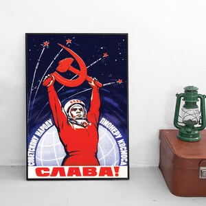 Puede incluir: Un póster enmarcado con una ilustración soviética de un cosmonauta con traje rojo sosteniendo una hoz y un martillo rojos contra un cielo azul oscuro estrellado. El texto incluye "СОВЕТСКОМУ НАРОДУ ПИОНЕРУ КОСМОСА" y "СЛАВА!" en letras rojas y blancas.