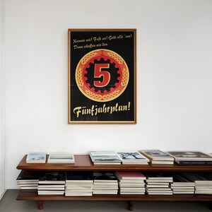K&ouml;nnte beinhalten: Ein gerahmtes rechteckiges Plakat mit schwarzem Hintergrund, das eine gro&szlig;e rote Zahl 5 in einem goldenen Zahnrad und &Auml;hrenkranz zeigt. Der deutsche Text lautet: Kommt mit! Fa&szlig;t an! Geht alle 'ran! Dann schaffen wir den F&uuml;nfjahrplan! Der Rahmen ist aus hellem Holz.