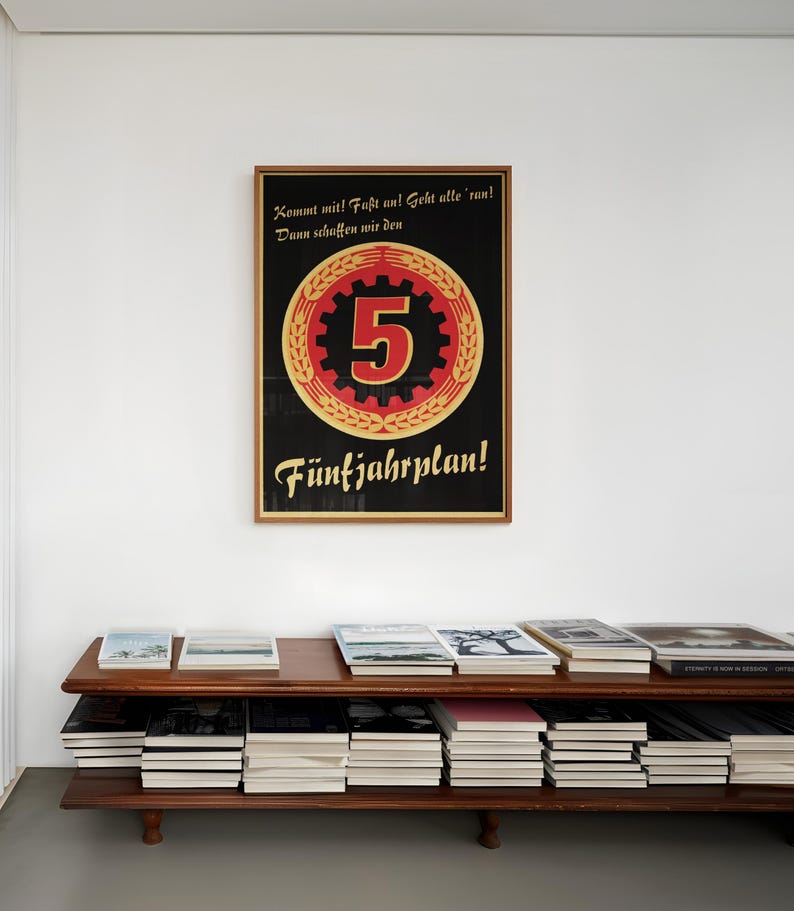 K&ouml;nnte beinhalten: Ein gerahmtes rechteckiges Plakat mit schwarzem Hintergrund, das eine gro&szlig;e rote Zahl 5 in einem goldenen Zahnrad und &Auml;hrenkranz zeigt. Der deutsche Text lautet: Kommt mit! Fa&szlig;t an! Geht alle 'ran! Dann schaffen wir den F&uuml;nfjahrplan! Der Rahmen ist aus hellem Holz.