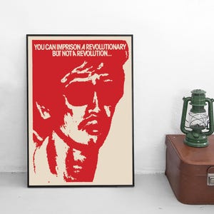 Può includere: Un poster incorniciato con una grafica rossa del volto di un uomo e il testo "YOU CAN IMPRISON A REVOLUTIONARY BUT NOT A REVOLUTION...". Il poster è su un muro bianco, accanto a una lanterna verde e una valigia marrone.