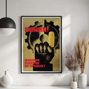 Pu&ograve; includere: Un poster incorniciato che mostra la sagoma nera di un pugno alzato davanti a un grande ingranaggio nero su sfondo giallo. Il testo in rosso recita "ПОМНИ!" in alto e "МАШИНА ОШИБОК НЕ ПРОЩАЕТ" in basso. Un dito giallo &egrave; puntato verso l'alto.