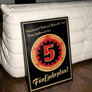 K&ouml;nnte beinhalten: Ein gerahmtes Poster mit schwarzem Hintergrund, das ein zentrales rot-schwarzes Zahnrad-Emblem mit einer gro&szlig;en Zahl 5 zeigt, umgeben von goldenen Weizen&auml;hren. Der Text lautet: Kommt mit! Fa&szlig;t an! Geht alle 'ran! Dann schaffen wir den F&uuml;nfjahrplan!