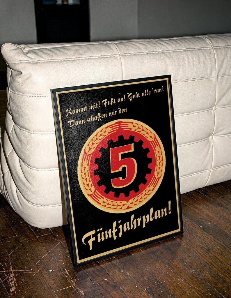 K&ouml;nnte beinhalten: Ein gerahmtes Poster mit schwarzem Hintergrund, das ein zentrales rot-schwarzes Zahnrad-Emblem mit einer gro&szlig;en Zahl 5 zeigt, umgeben von goldenen Weizen&auml;hren. Der Text lautet: Kommt mit! Fa&szlig;t an! Geht alle 'ran! Dann schaffen wir den F&uuml;nfjahrplan!
