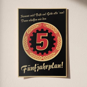 K&ouml;nnte beinhalten: Ein rechteckiges Plakat mit schwarzem Hintergrund, das in der Mitte ein rotes Zahnradsymbol mit einer gro&szlig;en Ziffer 5 zeigt, umgeben von einem goldenen &Auml;hrenkranz. Der Text lautet: Kommt mit! Fa&szlig;t an! Geht alle 'ran! Dann schaffen wir den F&uuml;nfjahrplan!