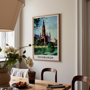 May include: A framed vertical wall art print featuring a colorful illustration of the Scott Monument in Edinburgh, Scotland. The artwork displays a tall gothic tower surrounded by green trees and parkland under a blue sky, with the text EDINBURGH below.