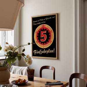 K&ouml;nnte beinhalten: Ein rechteckiges Wandplakat mit schwarzem Hintergrund, das ein zentrales rotes Zahnrad-Emblem mit einer gro&szlig;en Zahl 5 zeigt, umgeben von einem goldenen Weizenkranz. Der Text lautet: Kommt mit! Fa&szlig;t an! Geht alle 'ran! Dann schaffen wir den F&uuml;nfjahrplan!