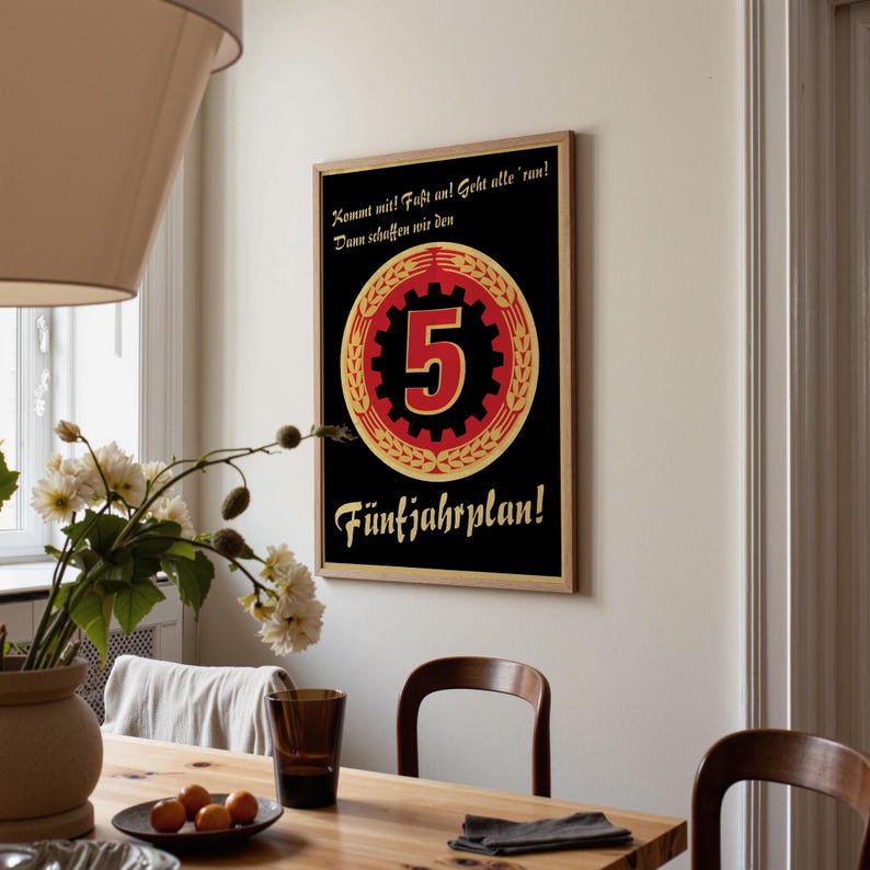 K&ouml;nnte beinhalten: Ein rechteckiges Wandplakat mit schwarzem Hintergrund, das ein zentrales rotes Zahnrad-Emblem mit einer gro&szlig;en Zahl 5 zeigt, umgeben von einem goldenen Weizenkranz. Der Text lautet: Kommt mit! Fa&szlig;t an! Geht alle 'ran! Dann schaffen wir den F&uuml;nfjahrplan!