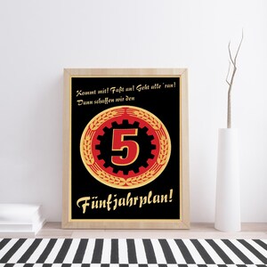 K&ouml;nnte beinhalten: Ein gerahmtes Poster mit schwarzem Hintergrund und einem zentralen roten Zahnrad-Symbol, das die Zahl 5 enth&auml;lt, umgeben von einem goldenen Weizenkranz. Der Text lautet: Kommt mit! Fa&szlig;t an! Geht alle 'ran! Dann schaffen wir den F&uuml;nfjahrplan! in Gold.