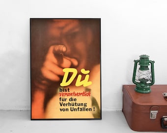 Op de afbeelding: Een ingelijste poster met een wazige afbeelding van een persoon die naar voren wijst met de gele tekst "Du" en de zwarte en rode tekst "bist verantwortlich für die Verhütung von Unfällen !" naast een groene lantaarn en een bruine koffer.