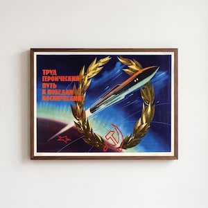 May include: A framed vintage-style poster featuring a dark blue space background with a rocket ascending through a golden laurel wreath. A red hammer and sickle symbol sits at the bottom. The text reads: ТРУД ГЕРОИЧЕСКИЙ ПУТЬ К ПОБЕДАМ КОСМИЧЕСКИМ.