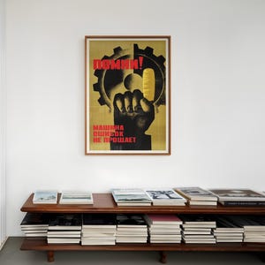 Pu&ograve; includere: Un poster incorniciato con sfondo giallo che presenta la sagoma di un ingranaggio nero e un pugno nero che tiene un oggetto giallo. Il testo in rosso recita "ПОМНИ!" in alto e "МАШИНА ОШИБОК НЕ ПРОЩАЕТ" in basso. Il poster &egrave; su una parete bianca.
