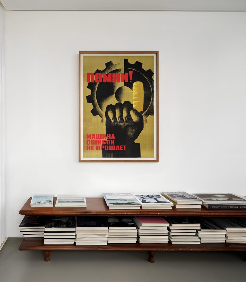 Pu&ograve; includere: Un poster incorniciato con sfondo giallo che presenta la sagoma di un ingranaggio nero e un pugno nero che tiene un oggetto giallo. Il testo in rosso recita "ПОМНИ!" in alto e "МАШИНА ОШИБОК НЕ ПРОЩАЕТ" in basso. Il poster &egrave; su una parete bianca.