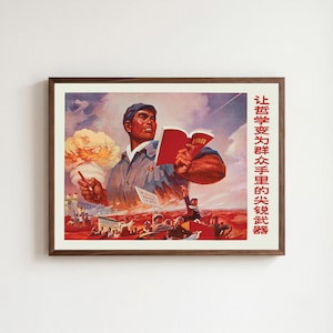 May include: A framed poster featuring a large illustration of a person in a blue cap and jacket holding a red book. In the background, a mushroom cloud rises above a crowd of people. Red Chinese text is printed vertically on the right side.