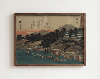 Op de afbeelding: Een ingelijste Japanse houtsnede met een landschap met traditionele huizen, een kasteel en figuren die over een weg reizen. De scène bevat gedempte tinten groen, bruin en oranje, met Japanse kalligrafie in het bovenste gedeelte.