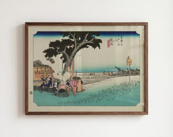 Op de afbeelding: Een ingelijste ukiyo-e houtsnede met een landschapsscène met een grote boom, figuren die rusten bij een bouwwerk en een verre horizon. Het kunstwerk bevat Japanse kalligrafie en een kleine vogel op een houten paal.