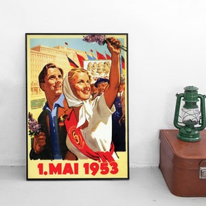 Könnte beinhalten: Ein gerahmtes Vintage-Propagandaplakat mit der Aufschrift 1.MAI 1953 zeigt ein lächelndes Paar mit lila Blumen. Die Frau trägt ein weißes Kopftuch und eine rote Schärpe mit der Zahl fünf. Im Hintergrund sind Flaggen und Porträts von Anführern zu sehen.