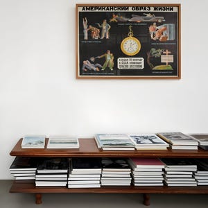 Może przedstawiać: Ramka z plakatem z tekstem "АМЕРИКАНСКИЙ ОБРАЗ ЖИЗНИ" (American Way of Life) w języku rosyjskim. Plakat przedstawia serię ilustracji przedstawiających r&oacute;żne aspekty amerykańskiego życia, w tym mężczyznę z bronią, samoch&oacute;d, zegar i krzyż. Plakat wisi na białej ścianie nad drewnianą p&oacute;łką z stosami książek.