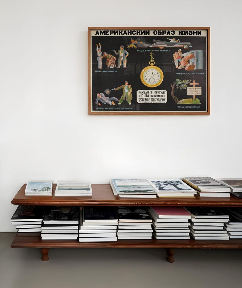 Może przedstawiać: Ramka z plakatem z tekstem "АМЕРИКАНСКИЙ ОБРАЗ ЖИЗНИ" (American Way of Life) w języku rosyjskim. Plakat przedstawia serię ilustracji przedstawiających r&oacute;żne aspekty amerykańskiego życia, w tym mężczyznę z bronią, samoch&oacute;d, zegar i krzyż. Plakat wisi na białej ścianie nad drewnianą p&oacute;łką z stosami książek.