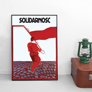 May include: A framed poster featuring the word SOLIDARNOŚĆ in black text at the top. The illustration depicts a person in red clothing walking across a grey cobblestone surface while carrying a large, flowing red flag against a white background.