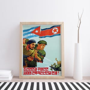 K&ouml;nnte beinhalten: Ein gerahmtes Propagandaposter mit zwei Soldaten in olivgr&uuml;nen Uniformen, die Gewehre halten und vor den Flaggen von Kuba und Nordkorea stehen. Darunter f&auml;llt eine Figur nach hinten. Das Poster enth&auml;lt unten roten koreanischen Text.