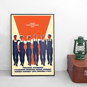 May include: A framed vintage Soviet propaganda poster featuring a group of people in work attire. The poster has text in Russian and a red and orange color scheme. A green lantern and a brown suitcase are next to the poster.