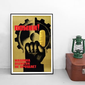 Pu&ograve; includere: Un poster incorniciato con la sagoma nera di un pugno alzato contro un grande ingranaggio su sfondo giallo. Il testo "ПОМНИ!" &egrave; scritto in rosso in alto e "МАШИНА ОШИБОК НЕ ПРОЩАЕТ" in rosso in basso. Una lanterna verde poggia su una valigia marrone.