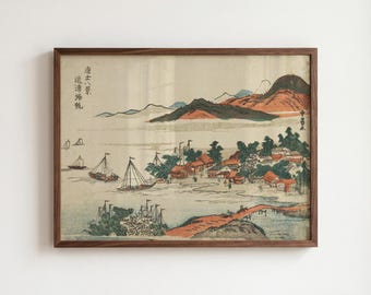 Op de afbeelding: Een ingelijste print met een landschap in Japanse houtsnijstijl met zeilboten op het water, een kustdorp met kleine huizen en verre bergen. Het kunstwerk bevat verticale zwarte tekstkarakters aan de linker- en rechterkant.