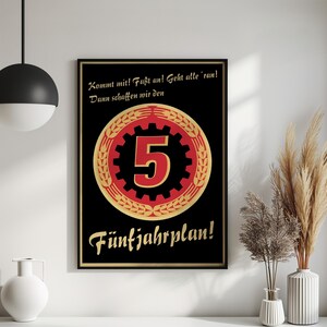 K&ouml;nnte beinhalten: Ein schwarzes rechteckiges Poster in einem d&uuml;nnen schwarzen Rahmen mit einem zentralen roten Zahnrad-Emblem und einer gro&szlig;en Zahl 5, umgeben von goldenen Weizen&auml;hren. Der Text lautet: Kommt mit! Fa&szlig;t an! Geht alle 'ran! Dann schaffen wir den F&uuml;nfjahrplan!