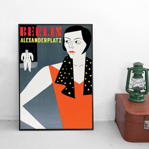 May include: A vintage poster with a black frame featuring a woman with short black hair wearing a red dress and a black and yellow polka dot scarf. The poster has the text "BERLIN ALEXANDERPLATZ" in red and black.